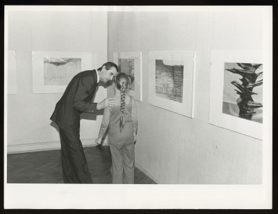 Czarno-białe zdjęcie. Duża grupa ludzi w przestrzeni wystawy. Na środku dwóch mężczyzn w garniturach ściska sobie dłonie.