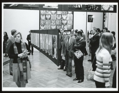 Czarno-białe zdjęcie. Na wystawie kilku zwiedzających, w tym na pierwszym planie starsi: mężczyzna i kobieta. Nad ich głowami wiszą zdjęcia.