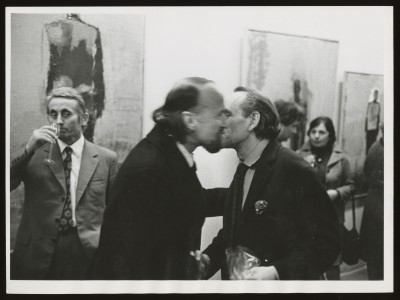 Czarno-białe zdjęcie. Dwóch mężczyzn całuje się w policzek, jeden jest rozmazany. Po lewej mężczyzna z kieliszkiem przy ustach, w tle wystawa.