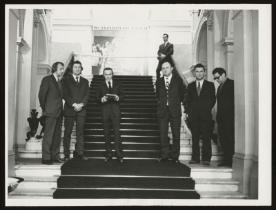 Czarno-białe zdjęcie. Mężczyźni na schodach: trzech z prawej, trzech z lewej strony. Wyżej mężczyzna przy rozpiętej wstędze, w tle duże zdjęcie.