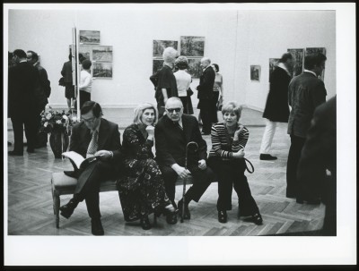 Czarno-białe zdjęcie. Mężczyzna ubrany na czarno podpisuje się w katalogu wystawy. W tle niewyraźny widok wystawy: obrazy na ścianie.