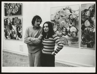 Czarno-białe zdjęcie. Mężczyzna w szarym swetrze i kobieta w swetrze w paski stoją na tle obrazów na białej ścianie.