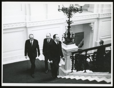 Czarno-białe zdjęcie. Wnętrze Zachęty. Na schodach pokrytych dywanem stoi mężczyzna. Poniżej trzy osoby, rozmawiają o czymś.