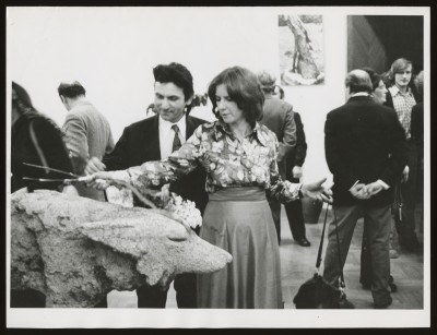 Czarno-białe zdjęcie. W centrum mężczyzna i kobieta. Kobieta zdaje się kłaść cięte kwiaty na kamiennej rzeźbie wilka. W tle wystawa i zwiedzający.
