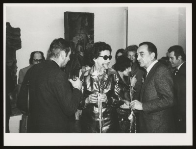 Czarno-białe zdjęcie. Kilka osób w sali wystawowej. Wśród nich kobieta w ciemnych okularach z kilkoma ciętymi kwiatami w dłoni. W tle wystawa.
