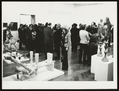 Czarno-białe zdjęcie. Kilka osób w sali wystawowej. Wśród nich kobieta w ciemnych okularach z kilkoma ciętymi kwiatami w dłoni. W tle wystawa.