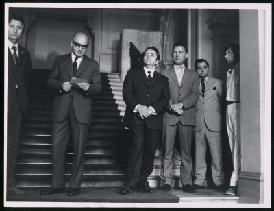 Czarno-białe zdjęcie. Schody Zachęty, na nich pięciu mężczyzn w garniturach, jeden w jasnej koszuli z krótkim rękawem. Mężczyzna pośrodku, w okularach, czyta z kartki. 