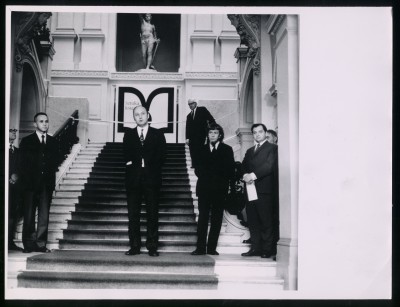 Czarno-białe zdjęcie. Kilku mężczyzn stojących na schodach Zachęty. W tle piąty mężczyzna przy rozciągniętej wstędze, dalej rzeźba gladiatora.