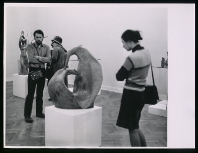 Czarno-białe zdjęcie. Grupa osób w przestrzeni wystawy. Przed nimi rzeźba na białym postumencie, za nimi rzeźby na szklanych postumentach.