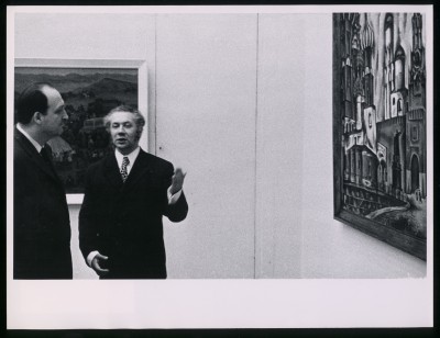 Czarno-białe zdjęcie. Trzech mężczyzn rozmawia ze sobą, są rozbawieni. W tle inni zwiedzający, dalej obrazy na ścianach.