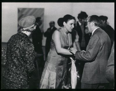 Czarno-białe zdjęcie. Po lewej kobieta w berecie stojąca tyłem. Po prawej mężczyzna i kobieta, trzymają się za dłonie jakby się witali.