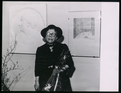 Czarno-białe zdjęcie. Na pierwszym planie ubrana na czarno kobieta w kapeluszu i okularach. Za nią po lewej płaskorzeźba i po prawej grafika. Zdjęcie 2: Czarno-białe zdjęcie. Grupa ludzi w tym kobieta z kwiatami, która stuka szklanką w kieliszek