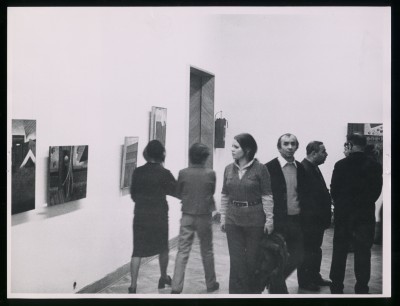 Czarno-białe zdjęcie. Na pierwszym planie nieostre postaci dwóch mężczyzn. W tle mężczyzna przy stole z kieliszkami, dalej obraz na ścianie.