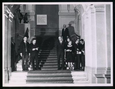 Czarno-białe zdjęcie. Dwie grupy: po lewej i prawej stronie schodów pokrytych długim dywanem na prawie całej szerokości. W tle wnęka z tablicą.