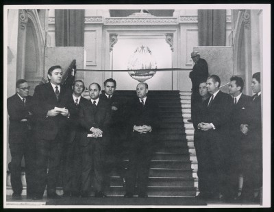 Czarno-białe zdjęcie. Grupa mężczyzn w garniturach stojących na wewnętrznych głównych schodach Zachęty. W tle w jasnej wnęce godło Rumunii.