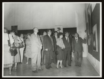 Czarno-białe zdjęcie. W przestrzeni wystawy tłum zwiedzających. Z prawej strony grupka mężczyzn w garniturach i jedna kobieta. Jeden z mężczyzn wskazuje na plakaty na ścianie. 