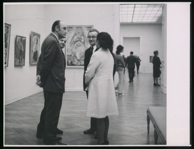 Czarno-białe zdjęcie. Cztery osoby rozmawiają ze sobą. W tle chodzą inni zwiedzający, na ścianach wiszą obrazy. Widoczne przejście do drugiej sali.