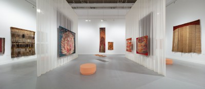 Fotografia kolorowa. Wnętrze sali wystawowej. Na ścianach i półprzeźroczystych kotarach, dzielących salę na trzy części, zawieszone ręcznie tkane tkaniny. Jedna podłużna tkanina umieszczona na przeźroczystej, falującej podstawie. Gobelin sprawia wrażenie j