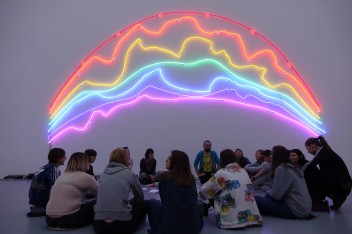 Na zdjęciu grupa młodzieży siedząca w kręgu w sali wystawowej. Na ścianie neon w kształcie tęczy.
