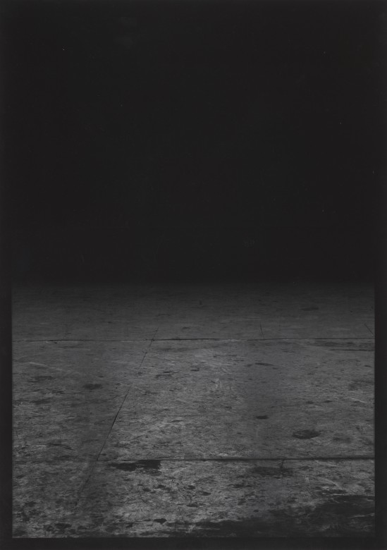 Czarno-biała fotografia w formacie pionowego prostokąta, niemal abstrakcyjna. Dolną połowę zajmuje ciemna podłoga, prawdopodobnie z płyt wiórowych. Górną połowę wypełnia czerń.