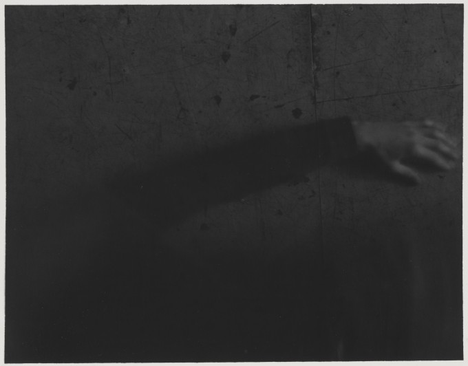 Czarno-biała fotografia w formacie poziomego prostokąta. Na ciemnoszarym tle płyty wiórowej ułożona poziomo ręka, widać lewą dłoń i długi czarny rękaw. W dolnej części kadru rozmyta ciemna plama.