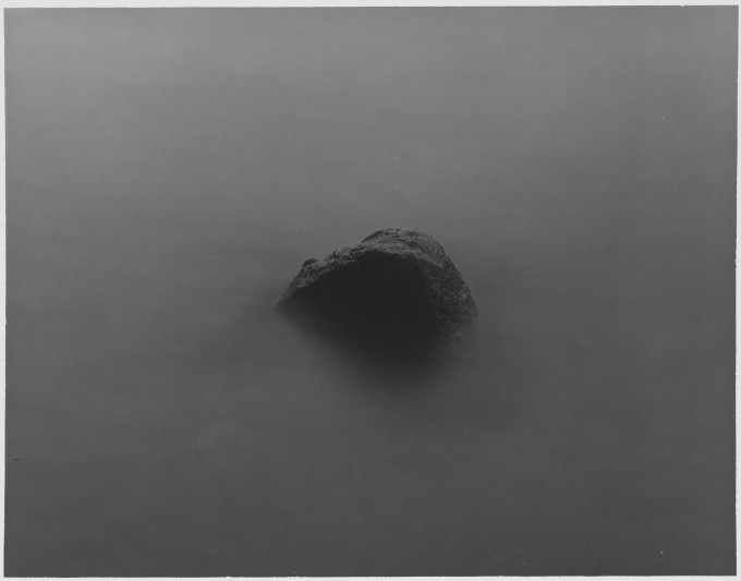 Czarno-biała fotografia w formacie poziomego prostokąta. Z jasnoszarej mgły wyłania się zaokrąglony szczyt skały w centrum kadru.