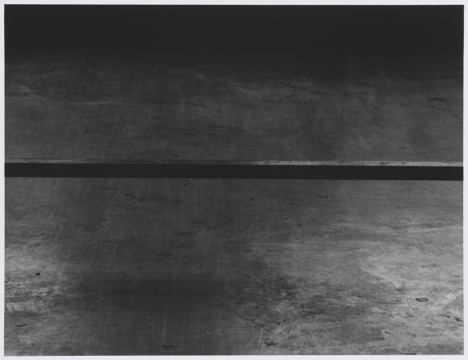 Czarno-biała fotografia w formacie poziomego prostokąta, niemal abstrakcyjna. W górnej i dolnej części kompozycji porysowane płyty, prawdopodobnie drewniane. Pomiędzy nimi w połowie kadru pozioma czarna luka.