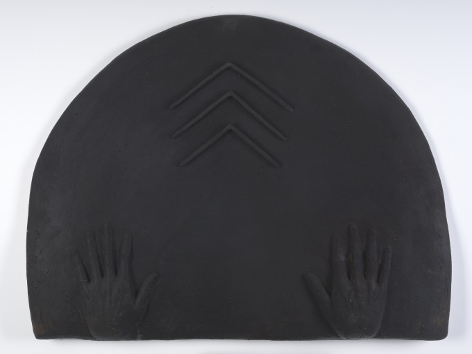 Płaskorzeźba z terakoty, wyglądem przypomina nieco matowy, prawie czarny metal. Płyta w formie półokręgu. Przy prostej dolnej krawędzi szeroko rozstawione realistyczne dłonie. Wyżej wypukły symbol w formie trzech "daszków" jeden nad drugim.
