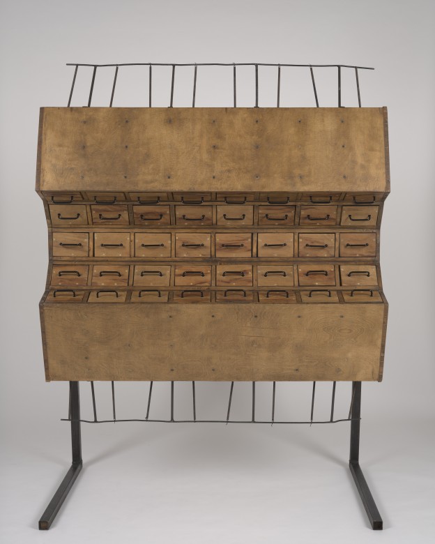Drewniana szafa z małymi szufladkami, wsparta na dwóch metalowych nogach. Przypomina katalog biblioteczny. Spodnia i górna część szafy są wygięte ku frontowi tak, że przekrój szafy przypomina kształtem półksiężyc. 