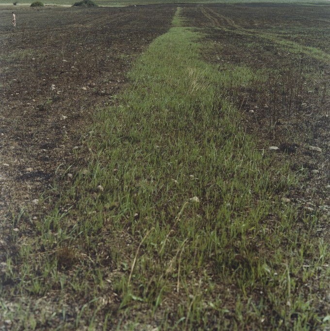 Fotografia o formacie kwadratu. Cały kadr wypełnia pole z widoczną brązową ziemią, miejscami porośnięte trawą. W oddali w lewym górnym rogu niewielka postać chodzącego po polu nagiego mężczyzny.