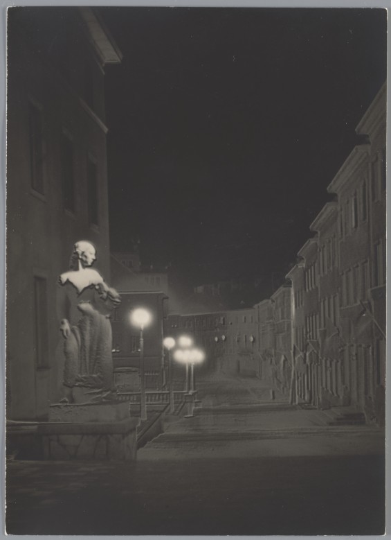 Zamglony widok uliczki nocą. Oświetlony latarniami chodnik i ściany kamienic. Na pierwszym planie biała rzeźba przedstawiająca kobietę.