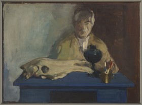 Wnętrze. Za niebieskim biurkiem siedzi starszy mężczyzna z siwymi wąsami. Wzrok ma zwrócony w prawą stronę. Na stole leży duża zwierzęca czaszka, obok stoi czarna lampa naftowa bez klosza, przed nią złote naczynie z dwoma długopisami w środku.