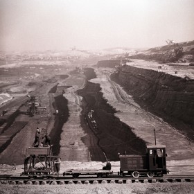 Na pierwszym planie lokomotywa na torowisku. W głębi ziemne tarasy, z których wydobywany jest piach lub żwir.