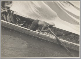 Zbliżenie na długą łódź z żaglem. W niej pochylony mężczyzna bez koszuli, który z wysiłkiem utrzymuje ster łodzi.