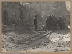 Tory kolejowe, po których kilku mężczyzn prowadzi nieduży wagon z gruzem. W tle ruiny, sterty desek i cegieł.