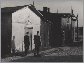 Dwóch mężczyzn idzie po brukowanej drodze. W tle fasada murowanego domu i drewniany dom.