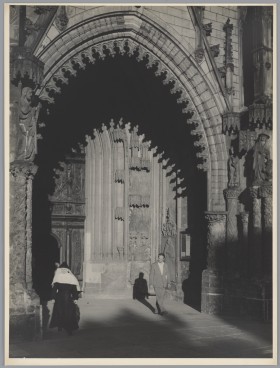 Wejście do katedry zakończone bogato zdobionym łukiem. Z wnętrza wychodzą siostra zakonna i mężczyzna w szarym garniturze.