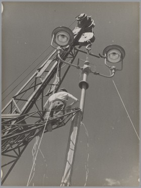 Wysoki dźwig trzyma podwójną metalową latarnię. Mężczyzna na dźwigu montuje ją do wysokiego betonowego słupa.