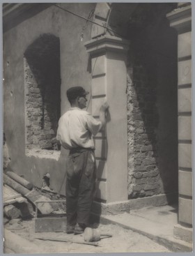 Przy odbudowywanej kamienicy stoi mężczyzna w białej koszuli, ciemnych spodniach i czapce. Układa na fragmencie ściany płaskie, kamienne płyty.