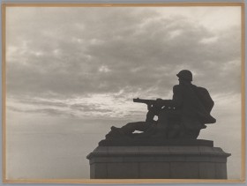 Ciemna figura żołnierza w hełmie z wycelowanym przed siebie karabinem. W tle zachmurzone niebo.