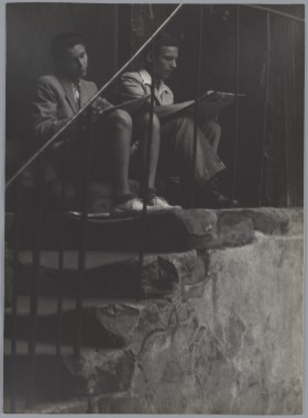 Kamienne schody z prosta balustradą. Na nich siedzi dwóch młodych mężczyzn. Rysują coś w skupieniu.